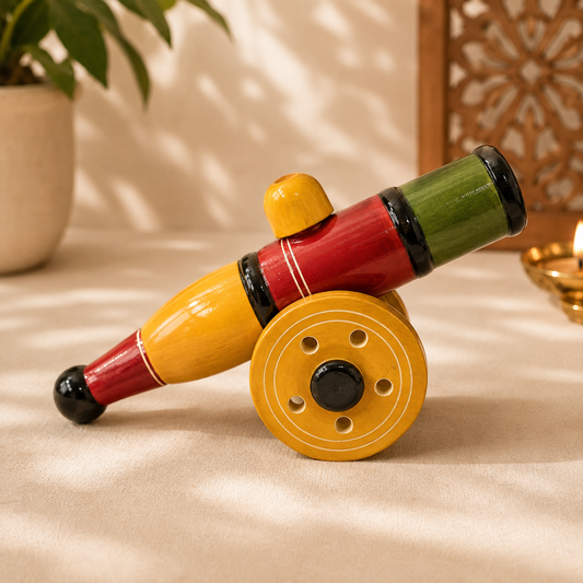 A hand-painted wooden toy cannon with yellow wheels, red and yellow barrels, and a green muzzle, displayed on a beige fabric surface with warm ambient lighting.